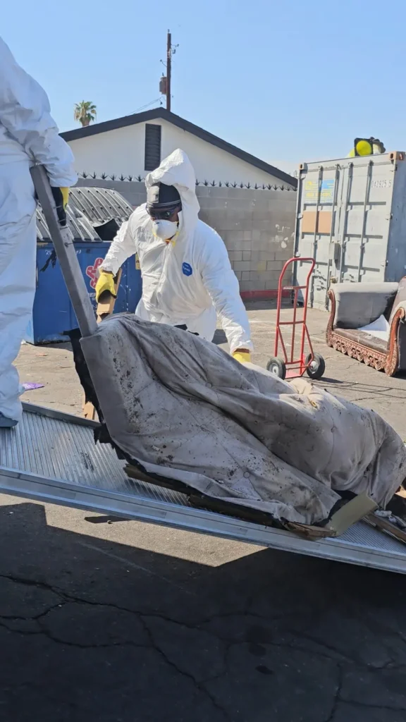 A person in a white protective suit loading a dirty mattress onto a cart.